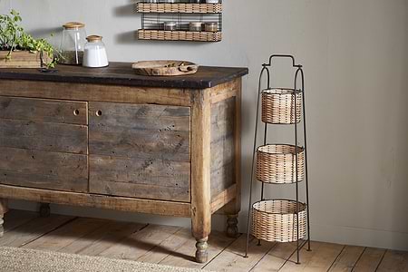 Against a light wall, jars and a wooden tray sit on a rustic sideboard. Next to it stands the nkuku Montali Iron & Rattan Three-Tiered Storage Stand—ideal for storing kitchen or pantry essentials in its handwoven baskets. The floor is wood.