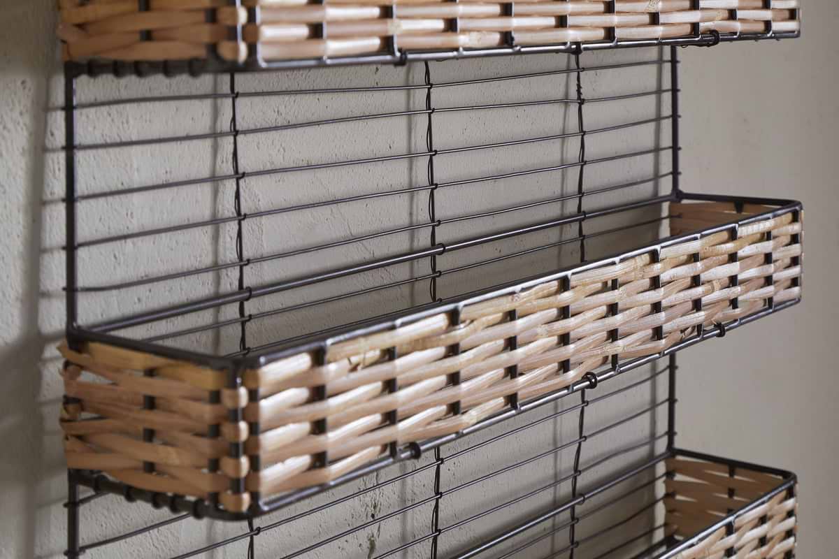 Close-up of the nkuku Montali Iron & Rattan Spice Rack - Natural, wall-mounted on a light grey textured wall. The rack has three empty rectangular wire baskets with handwoven rattan trim and black metal wire sides.