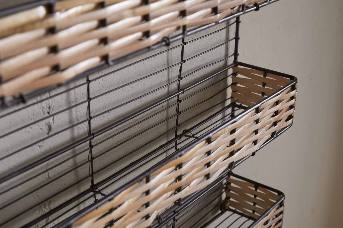 Close-up of nkuku's Montali Iron & Rattan Spice Rack - Natural, a wall-mounted organizer with three wire baskets and handwoven rattan fronts. The black frame contrasts with the light wall—ideal for kitchen storage or spices.