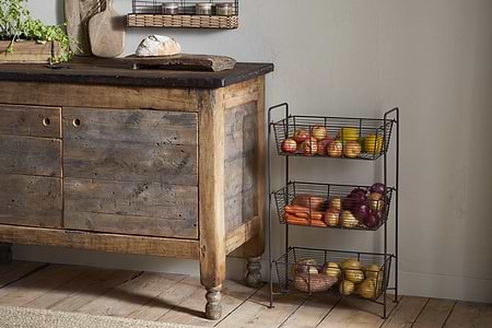 A rustic wooden cabinet sits on a wood floor beside the nkuku Inkollu Three Tier Storage Stand in antiqued brass, filled with apples, carrots, onions, and potatoes. A loaf of bread is on a cutting board atop the cabinet; kitchen items and greenery are visible.