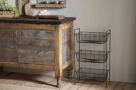 A rustic wooden sideboard with a dark countertop stands on a wooden floor. Next to it is the nkuku Inkollu Three Tier Storage Stand in antiqued brass. A loaf of bread and greenery rest on the sideboard, with a small shelf above.