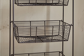 A close-up of the Inkollu Three Tier Storage Stand by nkuku in antiqued brass, featuring two empty rectangular wire baskets with a grid pattern—ideal for kitchen storage—set against a beige textured wall.