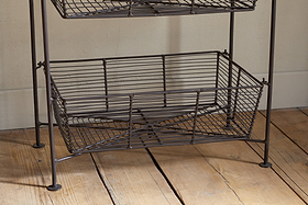 The empty Inkollu Three Tier Storage Stand by nkuku, featuring an antiqued brass metal frame and wire baskets, stands on a wooden floor with visible grain next to a cream wall and white baseboard.