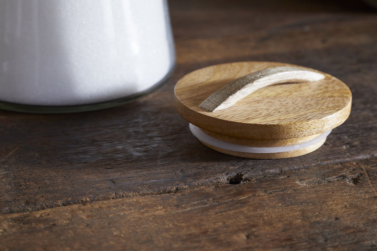 The nkuku Chaaval Storage Jar - Clear features a wooden lid with a curved handle and white rubber seal, resting on a rustic surface. Beside it, the glass jar is filled with white granules—ideal for stylish kitchen organisation.