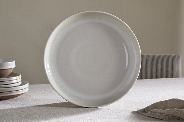 The Edo Dinner Plate - Terracotta by nkuku stands upright on a neutral tablecloth. Stacked everyday dinnerware sits to the left, with a striped chair and light wall creating a calm, minimalist dining scene.