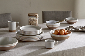 A beige tablecloth covers a dining table set with nkuku's Edo Dinner Plate in Terracotta, plus stacked ceramic bowls and mugs. A bowl of apricots, a jar with wooden lid, and a folded napkin complete the minimalist look.