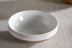 The nkuku Edo Cereal Bowl - Terracotta, featuring a glossy white interior and matte beige exterior, rests on a textured light fabric. Minimalist and empty, the bowl is gently lit by natural light, creating soft reflections on its surface.