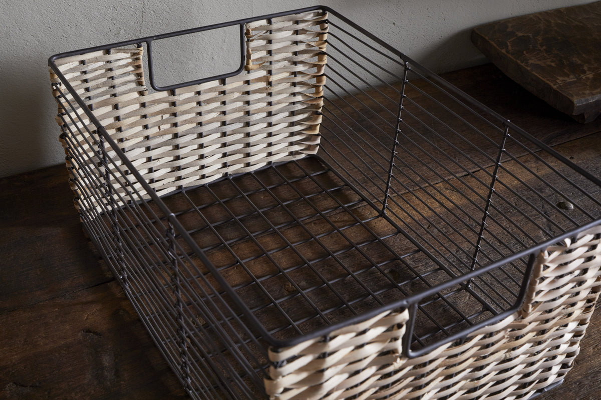 The Montali Iron & Rattan Storage Basket by nkuku, in natural white and brown with a metal frame, open handles, and grid-patterned base, rests on a dark wooden surface against a light wall.