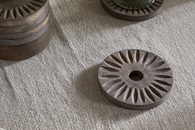 The Eisha Mango Wood Coasters by nkuku, in dark brown with hand-carved sunburst design, rest on textured beige fabric. A stacked set of six and a partially visible coaster share the elegant detailing.