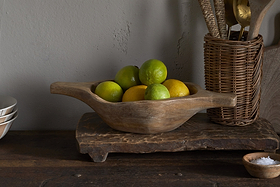The Naashta Handled Serving Bowl - Natural by nkuku, filled with green limes and yellow lemons, sits on a rustic board, flanked by wicker utensils in a basket and stacked dishes—perfectly arranged for a cozy kitchen display.