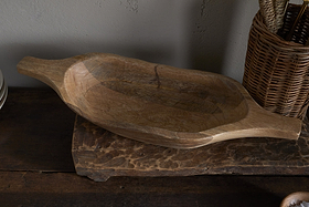 The Naashta Handled Serving Bowl - Natural by nkuku, an oval-shaped, hand-carved wooden bowl with handles, rests empty on a rustic kitchen surface. A woven utensil basket stands nearby against a neutral textured wall.