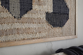 Close-up of the Kiran Large Textured Wall Art by nkuku, featuring natural and dark blue geometric handwoven fibers in a light wood frame, set against a cracked wall with a dark textured throw at the bottom.