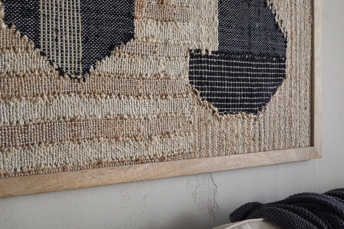 Close-up of the Kiran Large Textured Wall Art by nkuku, featuring natural and dark blue geometric handwoven fibers in a light wood frame, set against a cracked wall with a dark textured throw at the bottom.