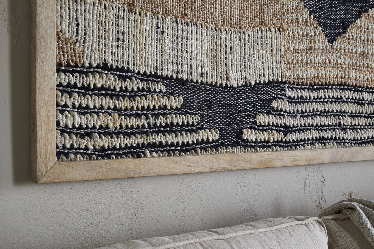 Close-up of nkuku's Birala Large Textured Wall Art in a wooden frame. The handwoven piece features cream, tan, and dark blue geometric patterns above a beige couch, with subtle wall imperfections adding character.