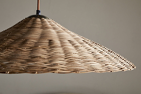 A close-up of the nkuku Sangu Rattan Pendant in Natural shows its large, conical wicker shade hanging from the ceiling. Soft light passes through the woven texture, casting gentle shadows on a neutral wall.