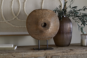 The Cala Reclaimed Wood Objet on Stand - Natural by nkuku, a round rustic sculpture with a center hole on a black metal stand, rests on a table beside a ribbed brown vase with branches, a candle, books, and circular artwork in the background.