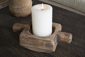 A lit white pillar candle sits in the nkuku Apu Reclaimed Wood Traditional Candle Holder - Natural, featuring two short wooden rod handles, set on a dark textured wooden surface. A blurred object appears in the background.