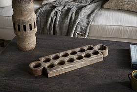 The Makala Reclaimed Wood Tealight Holder - Natural by nkuku, with carved circular pits, sits on a dark coffee table near a weathered accent, gray throw on a beige couch, and a black book titled THE LEGACY OF TITHES—a striking decor piece.