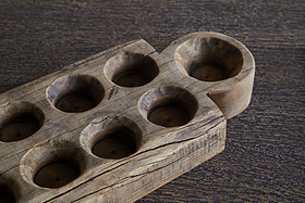 A close-up of the nkuku Makala Reclaimed Wood Tealight Holder - Natural, featuring eight round tealight slots and one larger semi-circular end slot. This decorative piece has a textured, weathered finish and rests unused on a dark, grainy surface.
