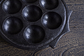 A close-up of the nkuku Maji Stone Multi Tealight Holder in Antique Black, featuring seven round wells, sits on textured wood. This functional décor piece has a seasoned, slightly worn surface for a distinctive look.