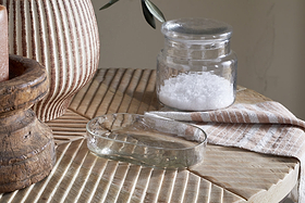 The Ilcoso Recycled Hammered Glass Soap Dish by nkuku rests on textured wood alongside a salt jar, striped cloth, ribbed ceramic vase, and rustic candle holder, bathed in soft natural light for a cozy, earthy bathroom feel.