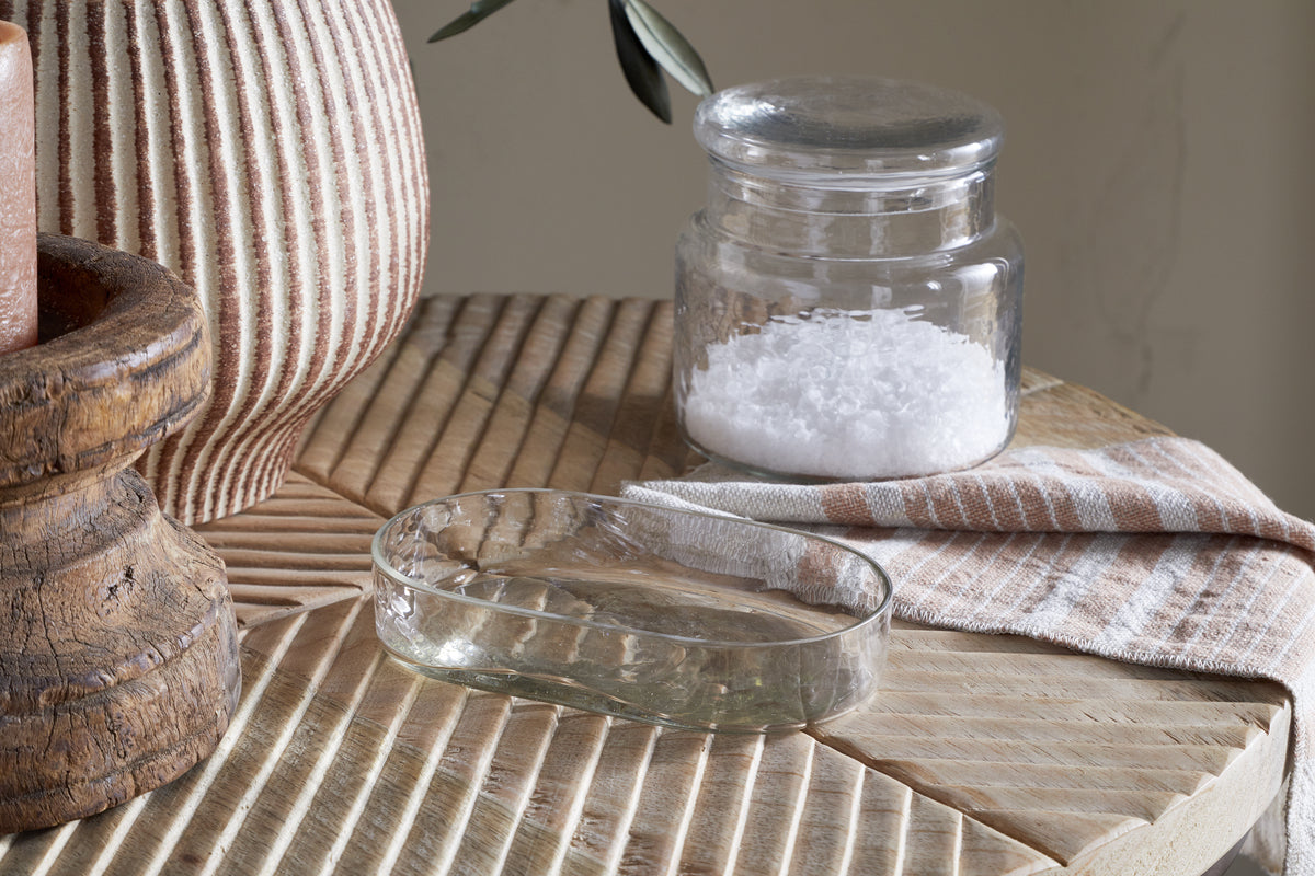 The Ilcoso Recycled Hammered Glass Soap Dish by nkuku rests on textured wood alongside a salt jar, striped cloth, ribbed ceramic vase, and rustic candle holder, bathed in soft natural light for a cozy, earthy bathroom feel.
