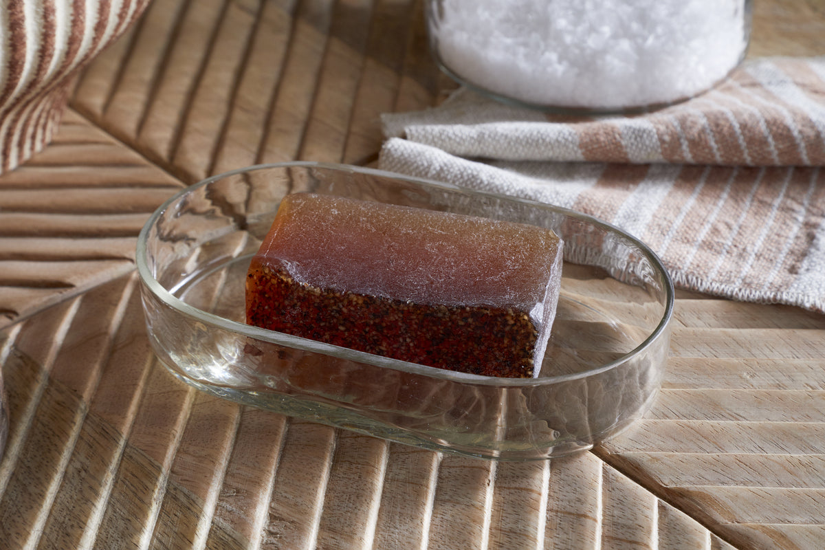 An amber-speckled soap rests in the Ilcoso Recycled Hammered Glass Soap Dish by nkuku on wood, with a striped cloth and a jar of coarse salt in the softly lit background, adding charm to the bathroom scene.