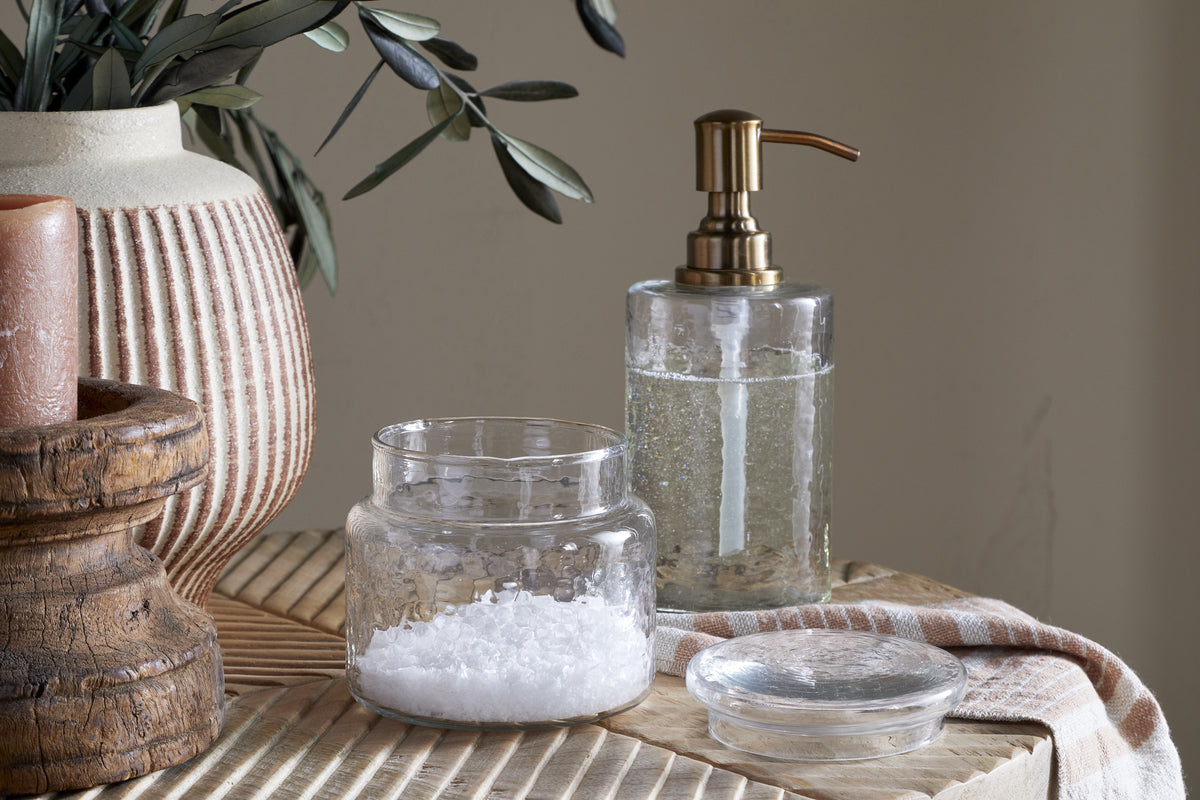 The nkuku Ilcoso Recycled Hammered Glass Storage Pot - Clear filled with white bath salts sits on a wooden surface next to its lid, accompanied by a glass soap dispenser, green leaves in a textured vase, and a brown candle in a wooden holder.