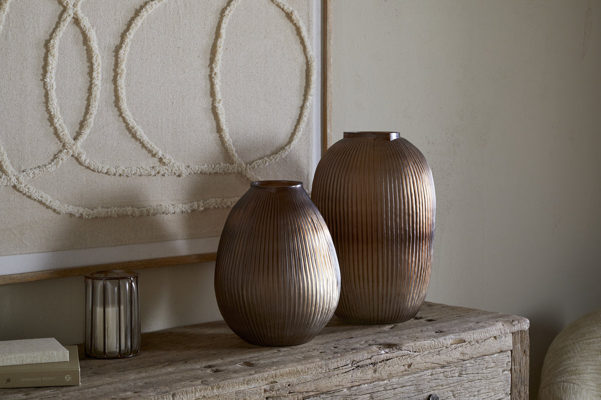 The nkuku Linsy Ribbed Glass Vase in Smoke Brown sits on a rustic wooden table by a candle and closed book, with neutral textile wall art featuring three large textured circles on a beige background behind it.