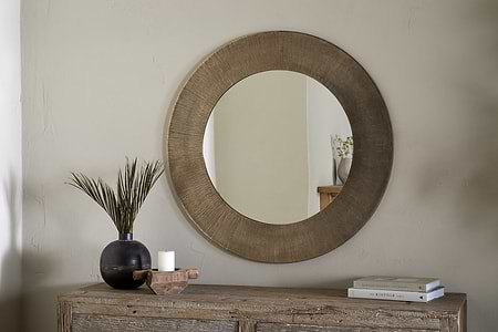 The nkuku Moto Round Mirror in Antique Brass hangs above a rustic wooden console table. On the table are a dark vase with tall green leaves, a white candle on a wooden tray, and two stacked books. The mirror reflects minimalist decor.