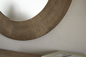 A close-up of the nkuku Moto Round Mirror in Antique Brass, featuring a textured frame and hanging on a light, subtly textured wall. A wooden surface with a closed book or box appears partially in the lower right corner.