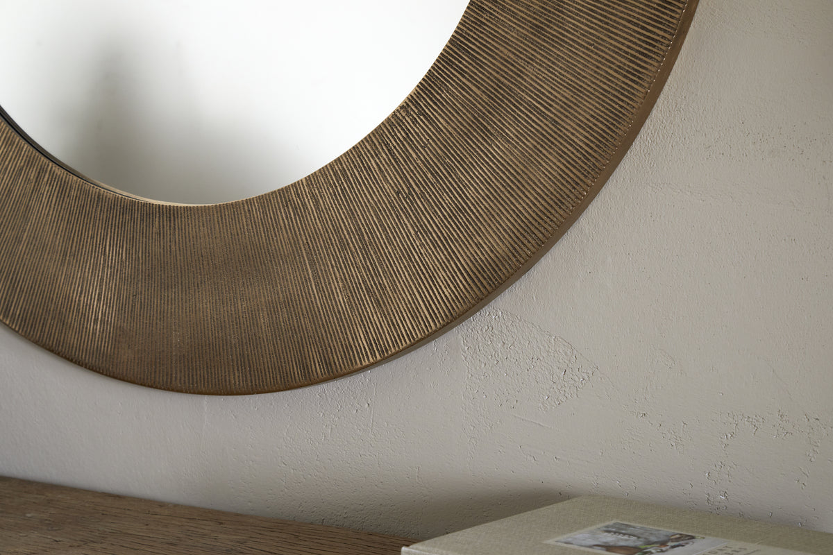 A close-up of the nkuku Moto Round Mirror in Antique Brass, featuring a textured frame and hanging on a light, subtly textured wall. A wooden surface with a closed book or box appears partially in the lower right corner.