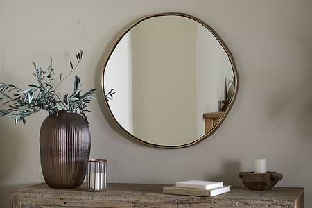 The Mchanga Organic Shape Mirror by nkuku, featuring an antique brass frame, hangs on a beige wall above a rustic wooden table with a ribbed brown vase of greenery, glass candle holder, stacked books, tray, and white candle.