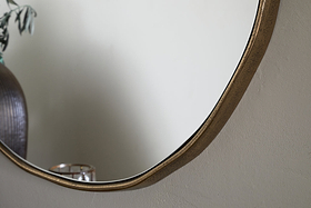 A close-up of the nkuku Mchanga Organic Shape Mirror in Antique Brass, hung on a light-textured wall. Its reflection shows part of a ribbed vase with greenery and a grid-pattern candle holder, both softly blurred.