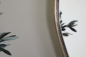 The nkuku Mchanga Organic Shape Mirror in antique brass is shown against a light wall, with green plant leaves visible in the foreground and reflected in the mirror, creating a soft, minimalist look.