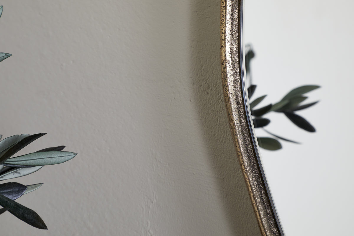 The nkuku Mchanga Organic Shape Mirror in antique brass is shown against a light wall, with green plant leaves visible in the foreground and reflected in the mirror, creating a soft, minimalist look.