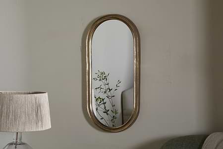 The Mbwa Oval Mirror in Antique Brass by nkuku hangs on a light gray wall. A tall lamp with a beige pleated shade stands to the left, while a light green sofa is partly visible on the right. The mirror reflects a green leafy plant.