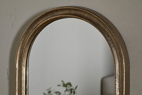 A close-up of the nkuku Mbwa Oval Mirror in Antique Brass, featuring a textured frame on a light gray wall. The mirror reflects blurred green plant leaves and part of neutral furniture.