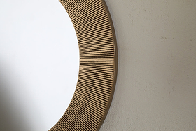 Close-up of the Putali Etched Mirror - Antique Brass by nkuku, featuring a sculptural, wide wooden frame with vertical grooves. The round mirror hangs on a light wall, reflecting a clean white space.
