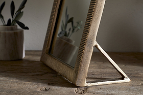 The nkuku Baja Standing Mirror in Antique Brass, featuring a textured frame and triangular stand, sits on a rustic wooden surface. Its reflection captures a blurred plant in a marble pot, softly lit by natural light.