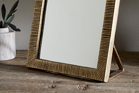 The nkuku Baja Standing Mirror in Antique Brass with a textured frame and stand is displayed on a rustic wooden surface, accompanied by a small marble vase with green stems to the left in a softly lit, minimal setting.
