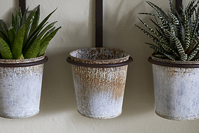 The nkuku Tukana Triple Wall Hung Planter - Zinc features three weathered metal pots; spiky green succulents fill the left and right, while the distressed middle planter is empty with visible rust marks.