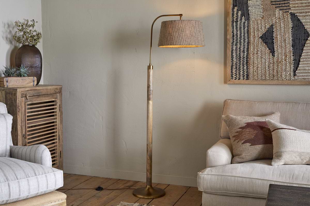 A cozy living room with the nkuku Kera Wood & Metal Floor Lamp in antique brass and natural beside a light sofa with patterned cushions, near a rustic mango wood base cabinet, abstract wall art, and a potted plant on the cabinet.