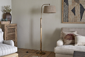 A cozy living room with a beige armchair, cream sofa with pillows, wooden furniture, plants, and a textured wall hanging. The space features the nkuku Kera Wood & Metal Floor Lamp in antique brass & natural mango wood.