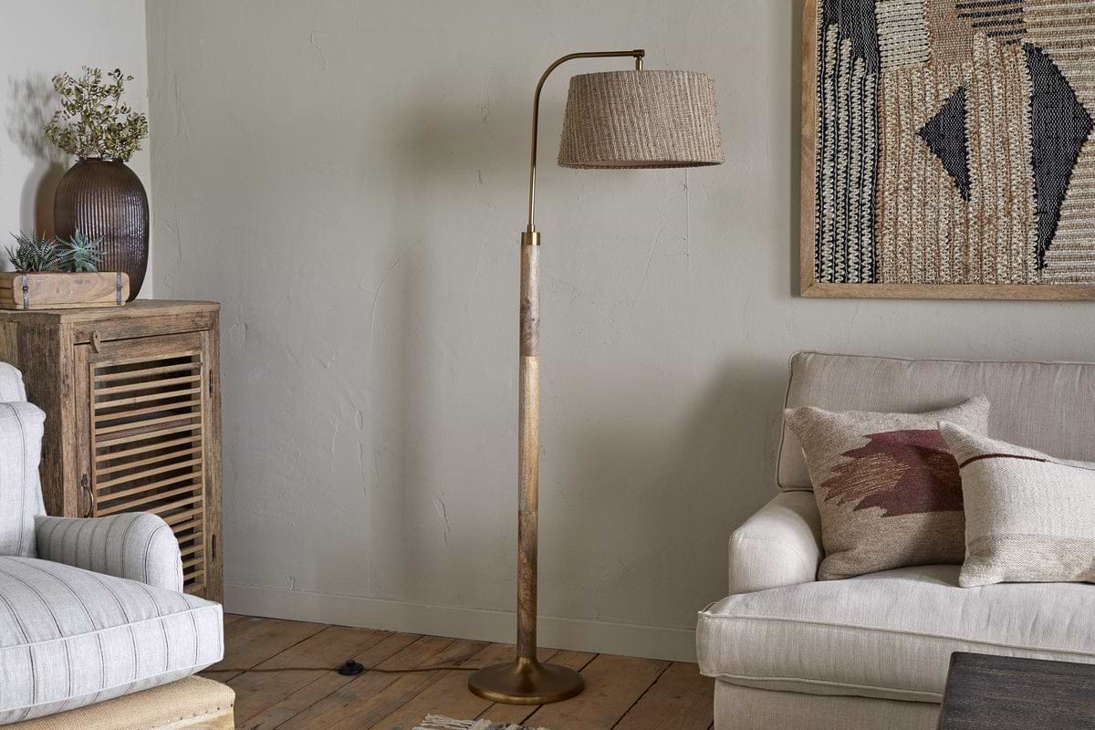 A cozy living room with a beige armchair, cream sofa with pillows, wooden furniture, plants, and a textured wall hanging. The space features the nkuku Kera Wood & Metal Floor Lamp in antique brass & natural mango wood.
