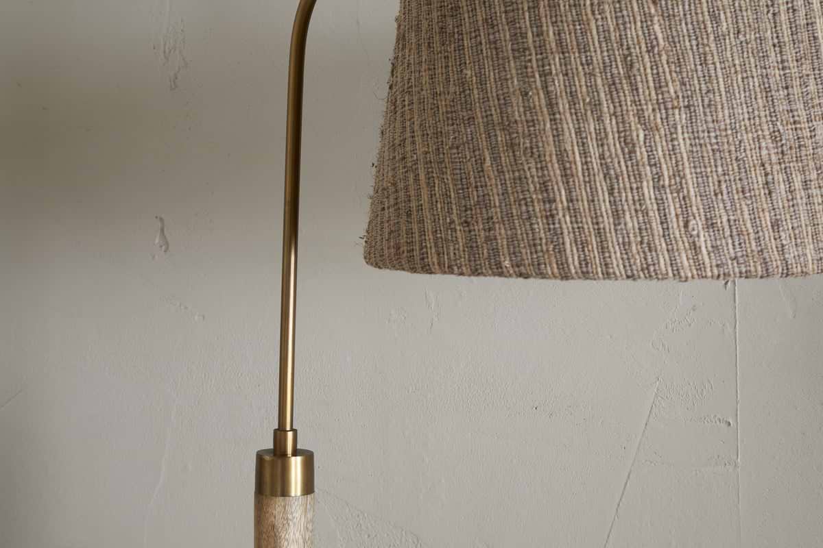 Close-up of the nkuku Kera Wood & Metal Floor Lamp in Antique Brass & Natural, showing part of its brass arm and textured beige shade against a light-colored, cracked wall.