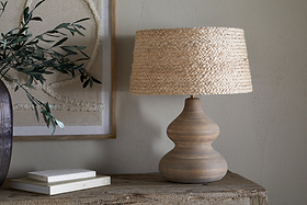 A wooden table displays the Dalat Ceramic Table Lamp - Natural by nkuku, featuring a wide, woven straw shade. Beside it are stacked books and a dark vase with green branches. Beige textured wall art decorates the light-colored wall behind.