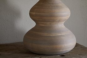 The Dalat Ceramic Table Lamp - Natural by nkuku features a smooth, light brown base with curvy, hourglass lines. Set on rustic wood against a neutral wall, its handcrafted look and subtle stripes are beautifully highlighted by natural light.
