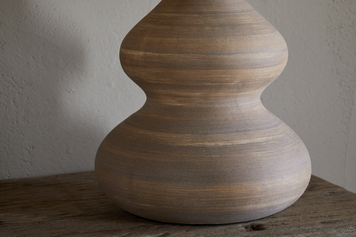 The Dalat Ceramic Table Lamp - Natural by nkuku features a smooth, light brown base with curvy, hourglass lines. Set on rustic wood against a neutral wall, its handcrafted look and subtle stripes are beautifully highlighted by natural light.