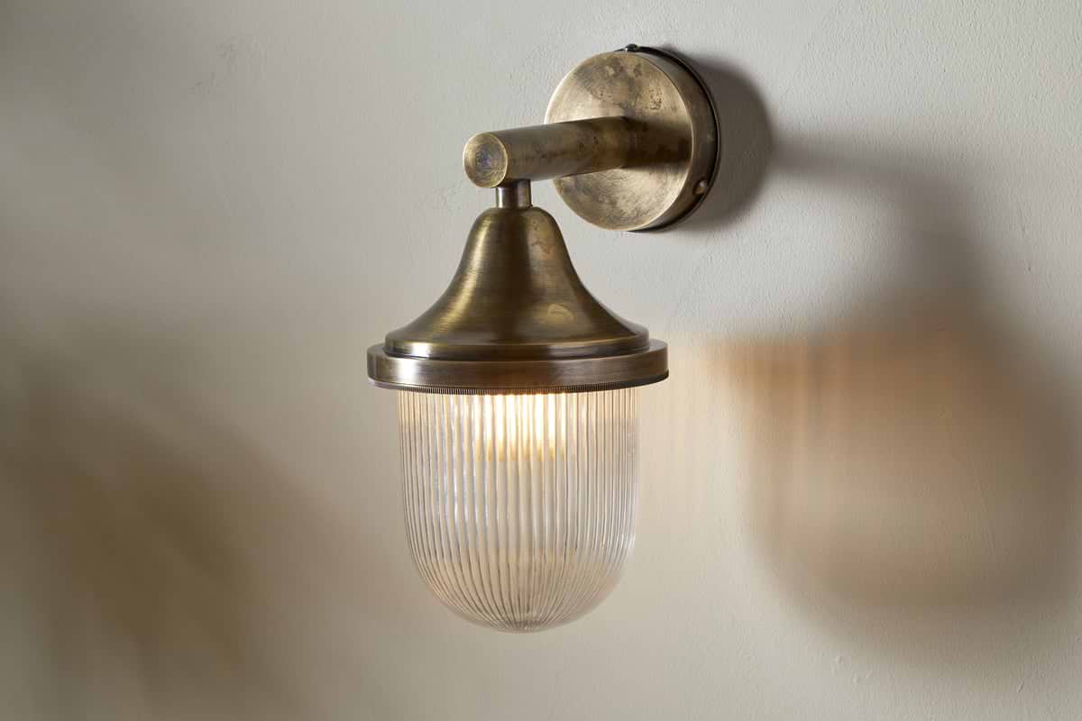 The Magola Outdoor Wall Lamp by nkuku features an antique brass finish and a cylindrical, ribbed glass shade. Mounted on a round base, it emits warm light, casting a soft glow and gentle shadows on light-colored walls.